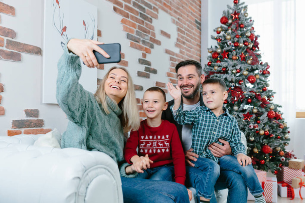 Beautiful happy married couple with two children using smart phone talking to friends or family congratulates Merry Christmas by video call conference app. Modern technology New Year 2022 congrats. - Foto, afbeelding