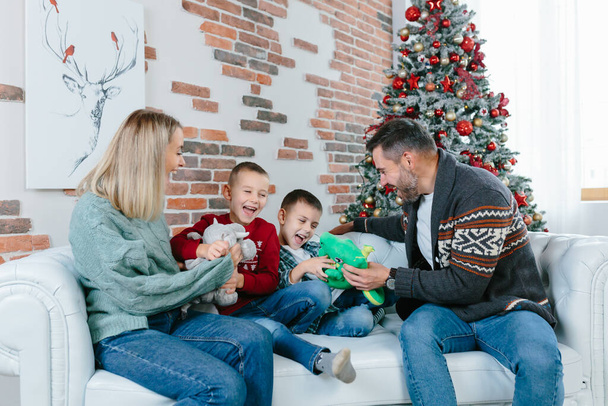 christmas and new year, young family of four men women husband and two little boys sons happily having fun together with toys having fun together sitting on sofa at home near Christmas tree - Foto, Imagen