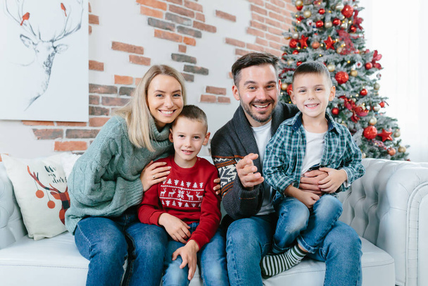 young family of four men woman husband and two little boys sons happily having fun together looking at webcam video communication with friends sitting on sofa at home near christmas and new year tree - Φωτογραφία, εικόνα