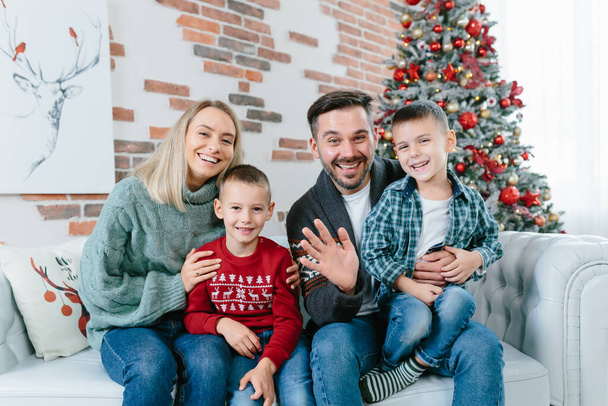 young family of four men woman husband and two little boys sons happily having fun together looking at webcam video communication with friends sitting on sofa at home near christmas and new year tree - Φωτογραφία, εικόνα