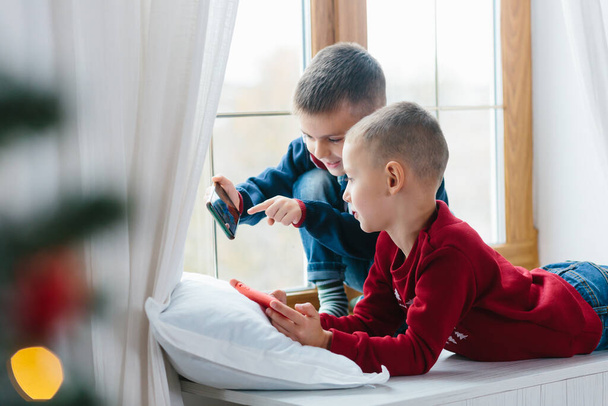 Two little boys kids playing together with mobile phones sitting on the windowsill of the apartment - Foto, Imagen