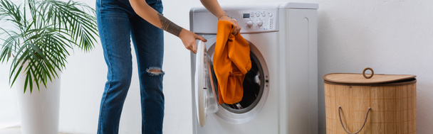 częściowy widok kobiety wkładającej ubrania do pralki w pobliżu kosza na pranie i roślin, baner - Zdjęcie, obraz