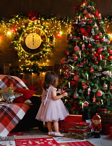 A little girl is decorating a Christmas tree with toys. Cute baby is getting ready to go home to celebrate Christmas. - Foto, Imagen