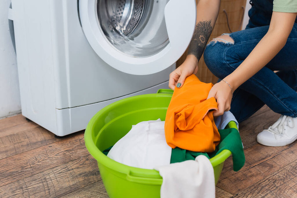cropped view of tattooed woman holding clothes near washer and laundry bowl - 写真・画像