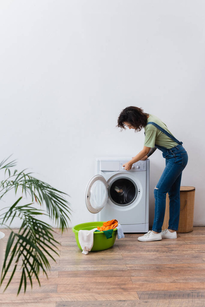 brunette woman operating washing machine near clothes in laundry bowl - Photo, Image