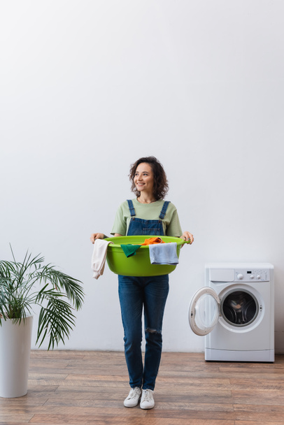 full length view of happy housewife holding laundry bowl with clothes near plant and washing machine - 写真・画像