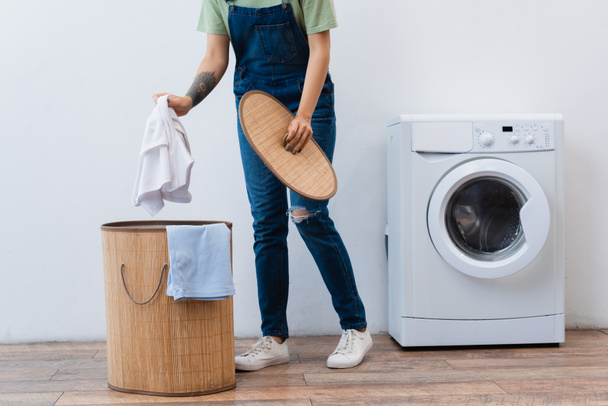 partial view of woman in denim overalls holding clothes near laundry basket and washing machine - Fotó, kép