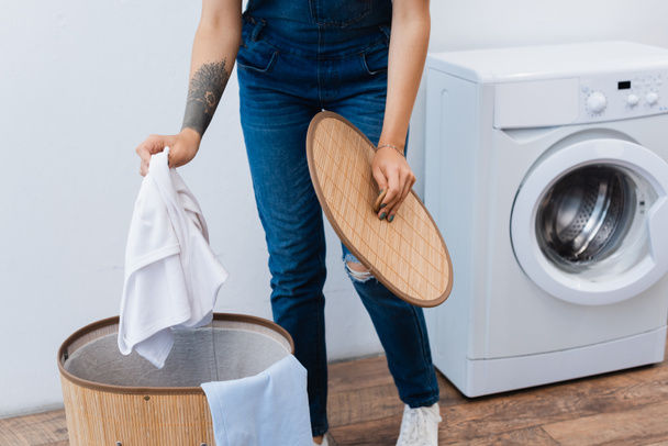 ořezaný pohled na tetovanou ženu držící oblečení v blízkosti prádelního koše a pračky - Fotografie, Obrázek