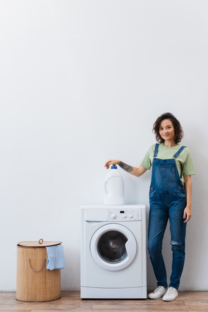 smiling housewife looking at camera near bottle of detergent on washing machine - Foto, Imagen