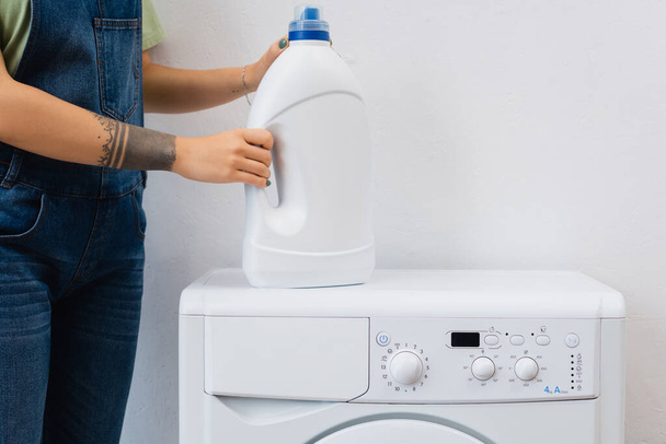 vista parcial de la mujer tatuada sosteniendo botella de detergente cerca de la lavadora - Foto, Imagen