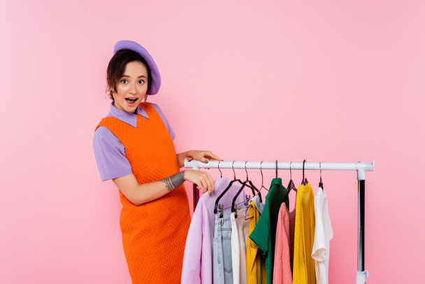 astonished woman in orange sleeveless dress looking at camera near rack with clothes isolated on pink - Foto, imagen