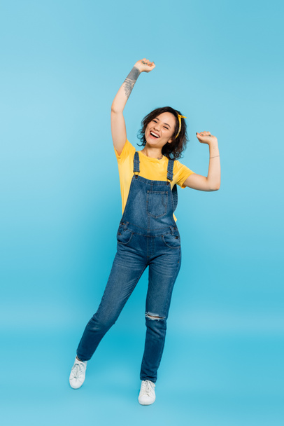 full length view of excited woman in denim overalls showing triumph gesture on blue - Фото, изображение
