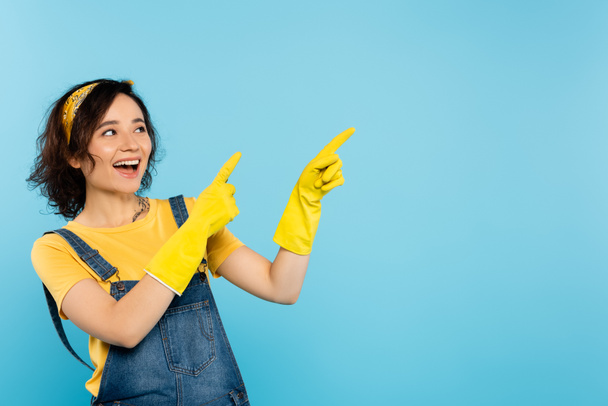 vzrušená žena ve žlutých gumových rukavicích dívá pryč a ukazuje prsty izolované na modré - Fotografie, Obrázek