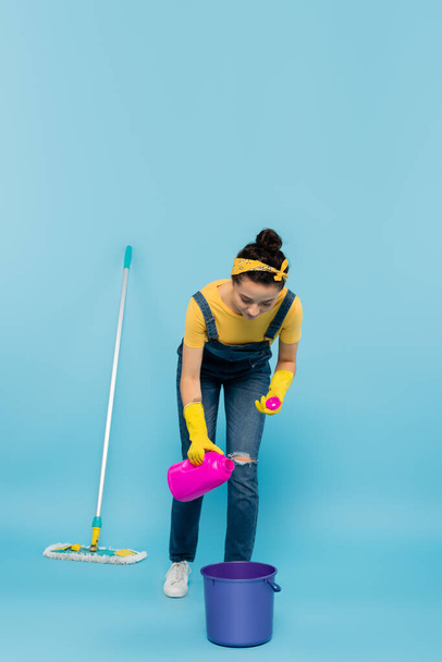 housewife in denim overalls and rubber gloves pouring detergent into bucket near mop on blue - Фото, изображение