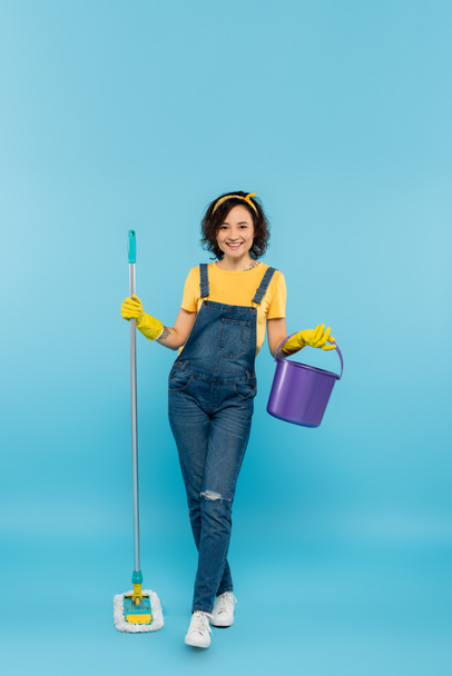 full length view of woman in yellow rubber gloves posing with bucket and mop on blue - Fotó, kép