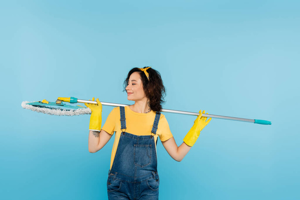 smiling woman in rubber gloves and denim overalls standing with mop isolated on blue - Фото, изображение