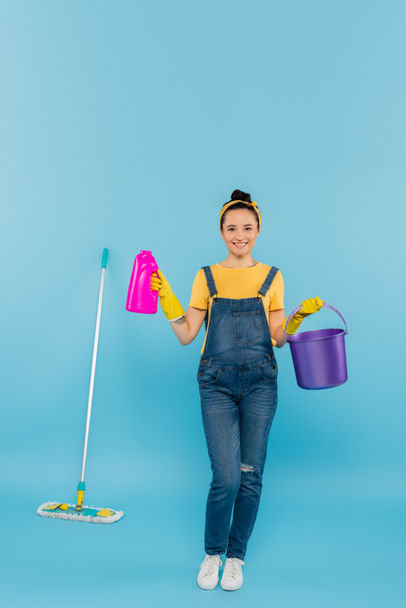 smiling housewife in denim overalls holding detergent and bucket near mop on blue - Foto, Bild