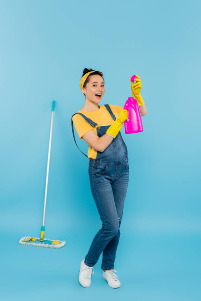 excited woman in denim overalls holding detergent while standing near mop on blue - Fotoğraf, Görsel