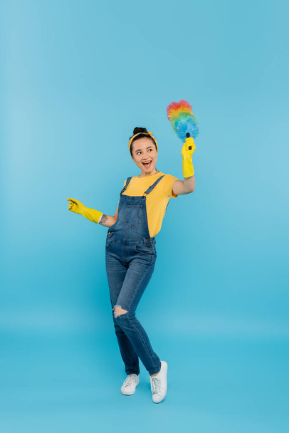 excited housewife in denim overalls waving dust brush while looking away on blue - Foto, imagen