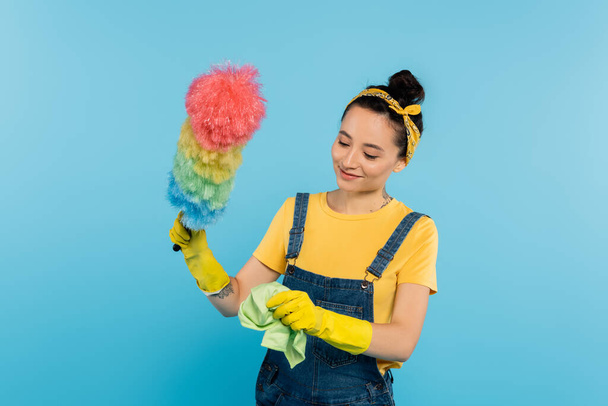smiling woman in denim overalls holding rag and colorful dust brush isolated on blue - Foto, afbeelding