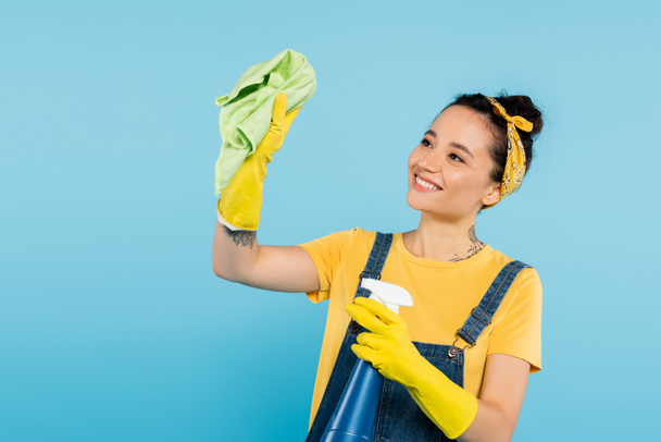 gelukkig vrouw in geel rubber handschoenen houden doek en spray fles met reinigingsmiddel geïsoleerd op blauw - Foto, afbeelding