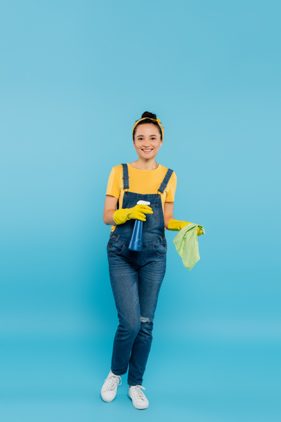 full length view of housewife in denim overalls and latex gloves holding rag and detergent on blue - Foto, afbeelding