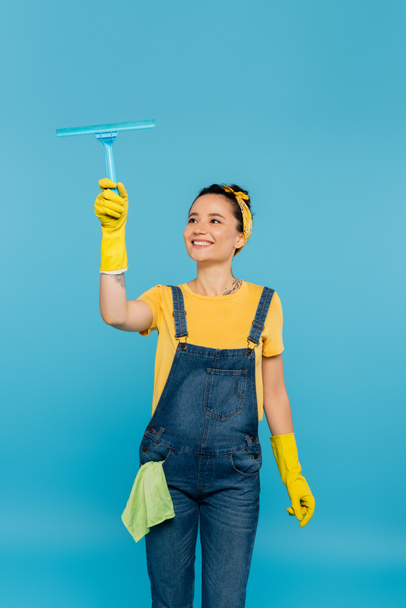 happy housewife with rag in pocket of denim overalls holding window cleaner isolated on blue - Fotó, kép