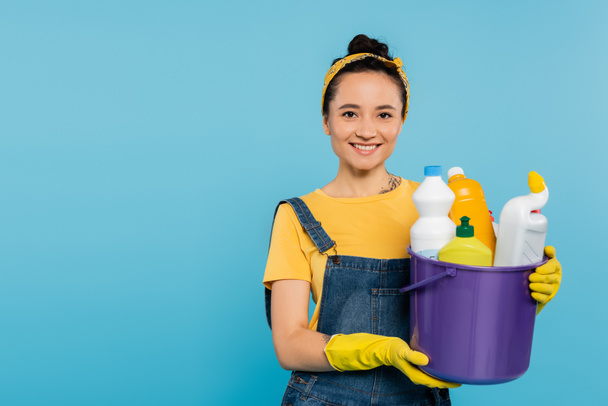 young housewife in yellow rubber gloves holding bucket with detergents isolated on blue - Photo, Image