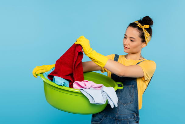 housewife in yellow rubber gloves looking at clothing in laundry bowl isolated on blue - Photo, Image
