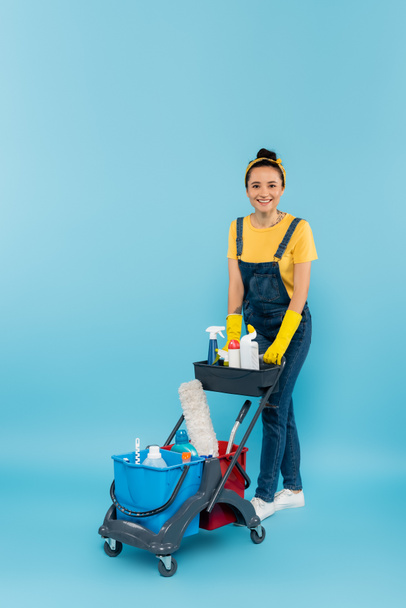 happy cleaner in denim overalls looking at camera near trolley with cleaning supplies on blue - Фото, изображение