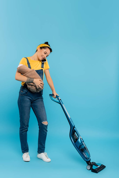happy woman with baby doll talking on mobile phone while vacuuming on blue - Foto, Imagem