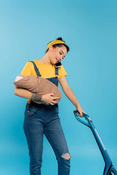 smiling housewife holding baby doll, talking on smartphone and vacuuming isolated on blue - Photo, Image