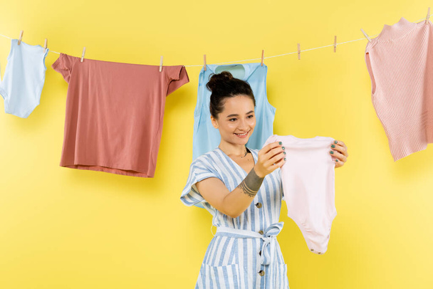 smiling housewife holding baby romper near laundry hanging on rope on yellow background - Photo, image