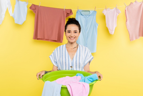 黄色の背景にロープでぶら下がった服の近くに洗濯器を持った幸せな主婦 - 写真・画像