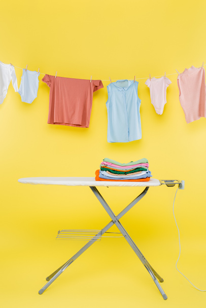 stack of clean clothes on ironing board under laundry hanging on rope on yellow background - Photo, Image