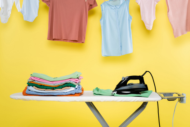 elektrisches Bügeleisen und Stapel sauberer Wäsche auf Bügelbrett unter Kleidung, die auf gelbem Hintergrund hängt - Foto, Bild