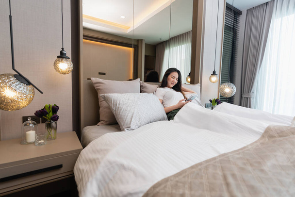 Slaapkamer concept een meisje met slaperig gevoel leunen haar rug op een aantal grote en comfortabele kussens op het bed kijken op het mobiele scherm voor sociale media. - Foto, afbeelding