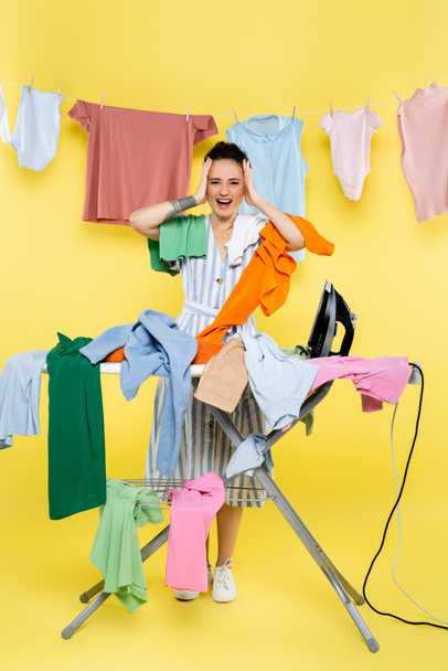 sorprendido ama de casa tocando la cabeza y gritando cerca de la ropa en tabla de planchar en amarillo - Foto, Imagen