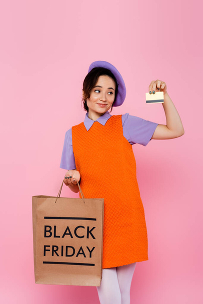 pleased girl in purple beret holding credit card and shopping bag with black friday lettering isolated on pink - Foto, Bild