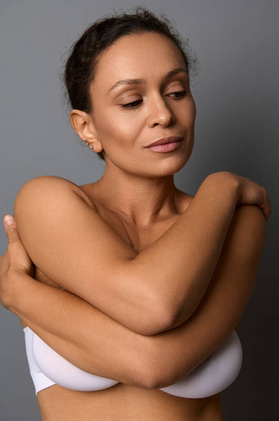 Close-up beauty portrait of self-confident middle aged African woman with perfect clean shiny healthy skin, hugging herself, isolated over gray background with copy space. Skin and body care concept - Photo, Image