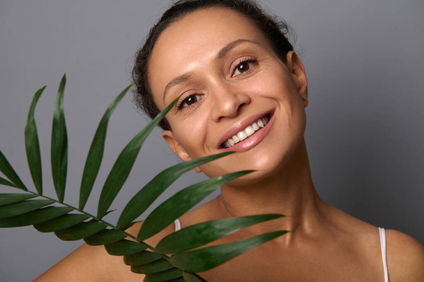 Aantrekkelijke vrouw met mooie glimlach en schone huid kijkt naar de camera met een palmblad geïsoleerd over grijze achtergrond met kopieerruimte. Spa, schoonheidsbehandeling, anti-aging, natuurlijke cosmetica concept - Foto, afbeelding