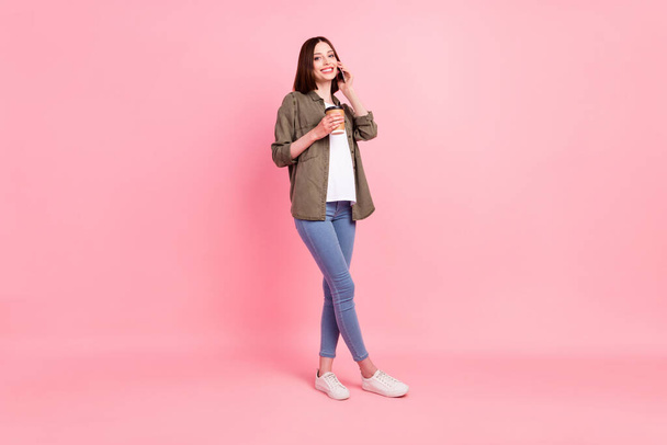 Full length photo of optimistic brunette hairdo lady talk telephone hold coffee wear green shirt jeans isolated on pink color background - Фото, зображення