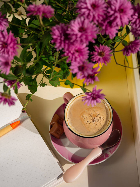 Kahve fincanı çikolatalı defter güzel pembe sonbahar kasımpatı çiçekleri pencerenin üzerinde sarı demlikte. Sıcak ev titreşimleri moda oluyor. Kentsel alanlar bitkiler soğuk havada sıcak kakao içiyor. Günaydın konsept. - Fotoğraf, Görsel