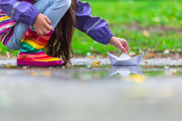 Щаслива дівчинка з різнокольоровими гумовими черевиками, що граються з паперовим човном у калюжі восени на природі
 - Фото, зображення