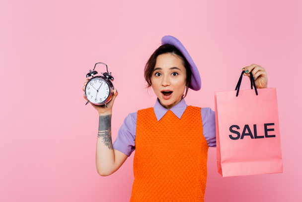 ヴィンテージ目覚まし時計を持つ明るい服の興奮した女性とピンクの上に隔離された販売ショッピングバッグ - 写真・画像