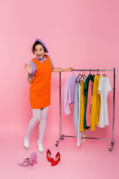 cheerful woman waving hand near footwear and rack with clothes on pink - Фото, изображение