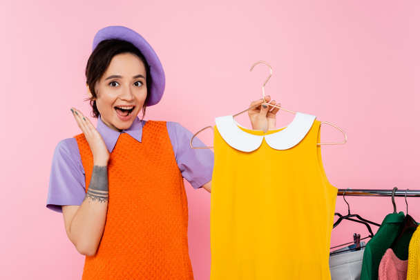amazed woman showing wow gesture near yellow sleeveless dress and rack with clothes isolated on pink - Zdjęcie, obraz