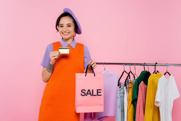 stylische Frau mit Kreditkarte und Einkaufstasche mit Verkauf Schriftzug lächelnd in der Nähe Kleidungsstücke isoliert auf rosa - Foto, Bild