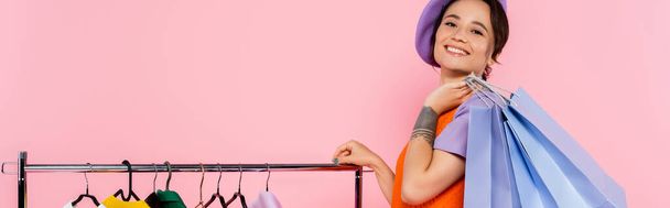 joyful woman in purple beret holding shopping bags near rack with garments isolated on pink, banner - Fotoğraf, Görsel