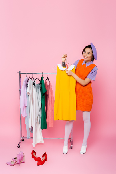 happy woman holding yellow sleeveless dress with sale tag near footwear and rack with garments on pink - Foto, Imagem